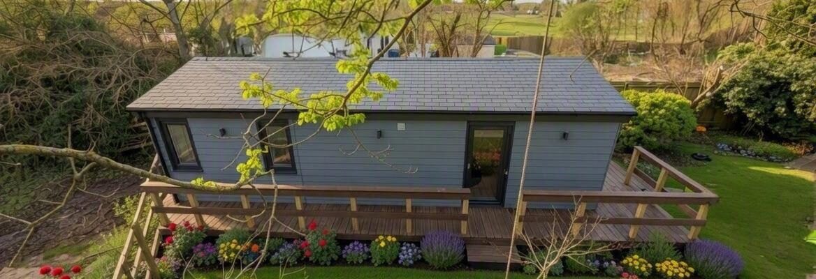 Dark grey Cedral clad one-bedroom garden annexe with a Tapco slate-tiled roof, set on an elevated timber deck with a wooden railing, surrounded by flowering borders and mature trees.